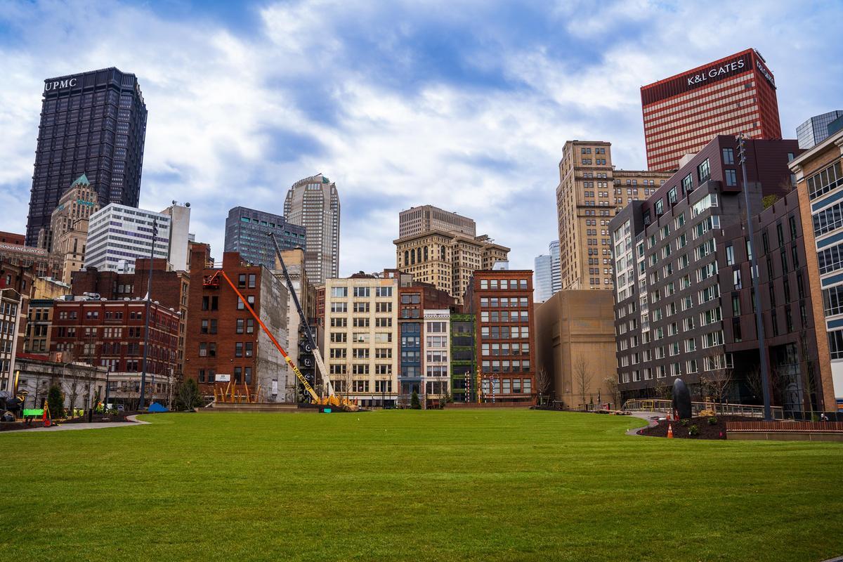 A city park with a large green lawn is surrounded by modern and historic buildings under a partly cloudy sky. Construction equipment is visible near the buildings.
