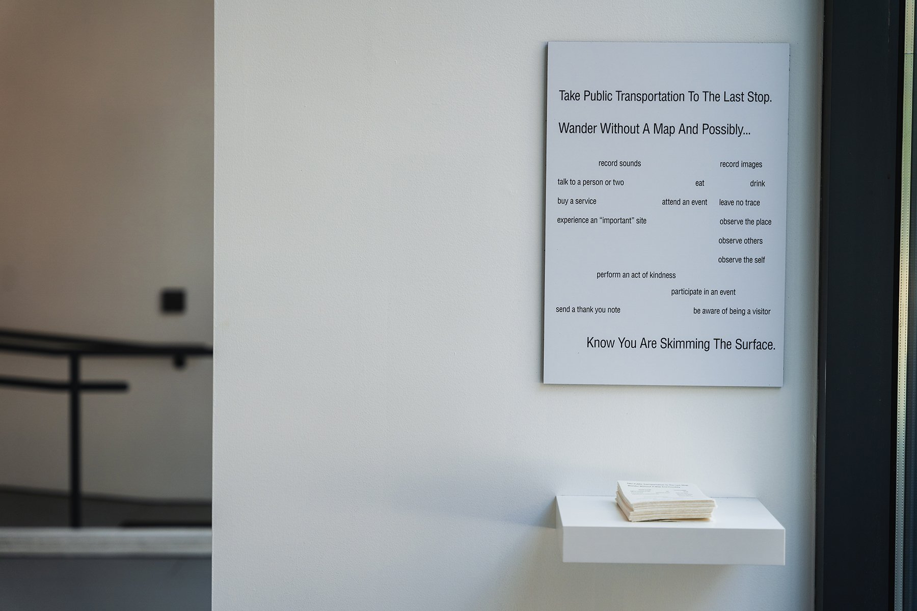 a poem on a white gallery wall. a small ledge below holds a stack of small printed versions of the same poem.