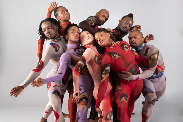 A group of dancers cluster together in multi-colored leotards and colorful makeup.