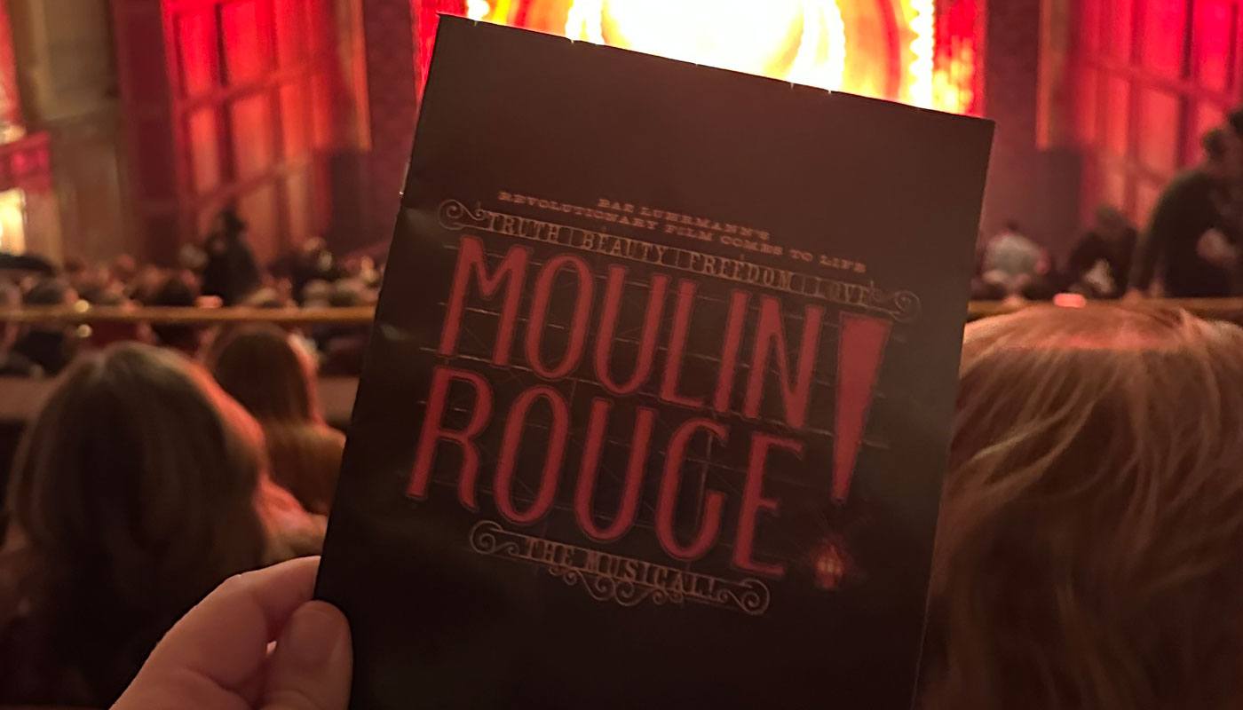 a hand holds up a Moulin Rouge program book with the Benedum stage in the background