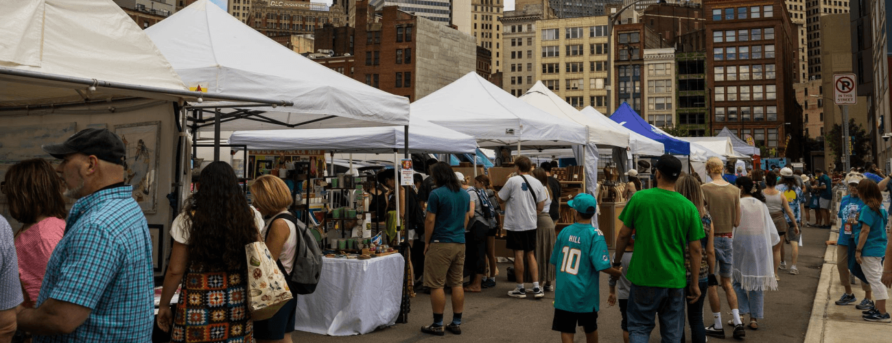 A crowd mills around a line of artist booths with Pittsburgh city buildings in the background.