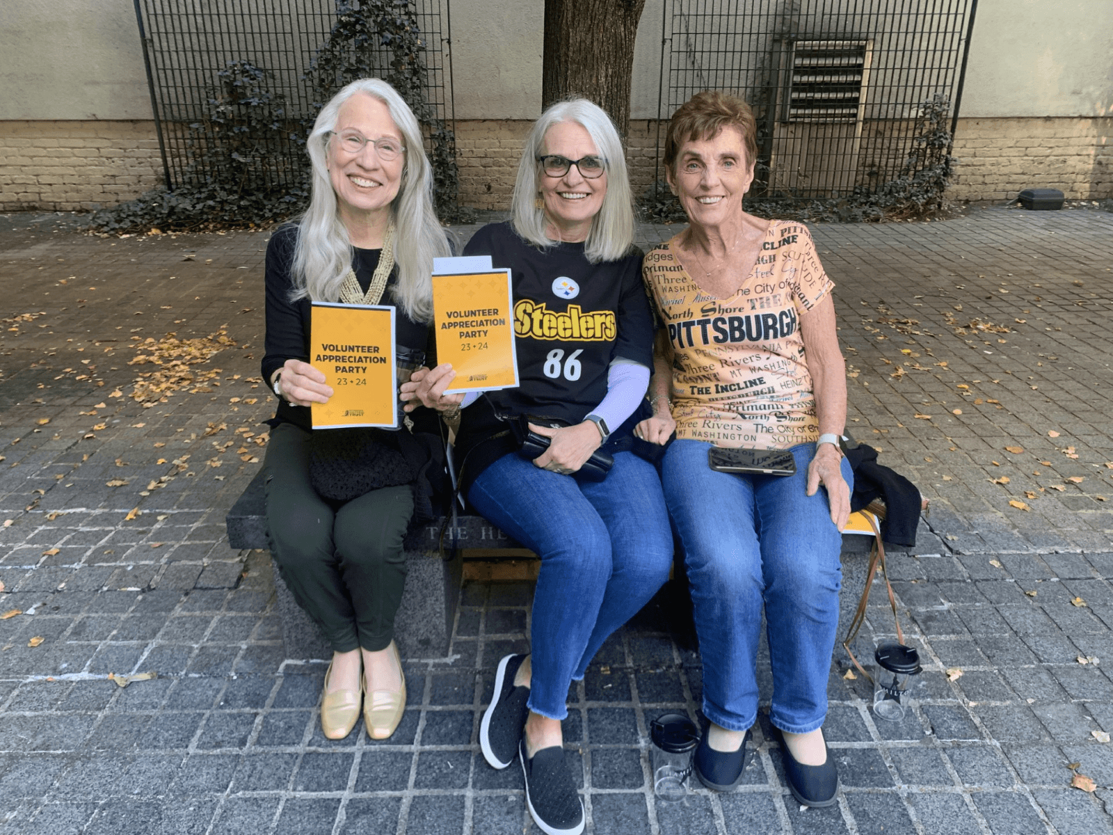 three volunteers wearing Pittsburgh and Steelers gear hold up their black and gold Volunteer Appreciation Party programs.