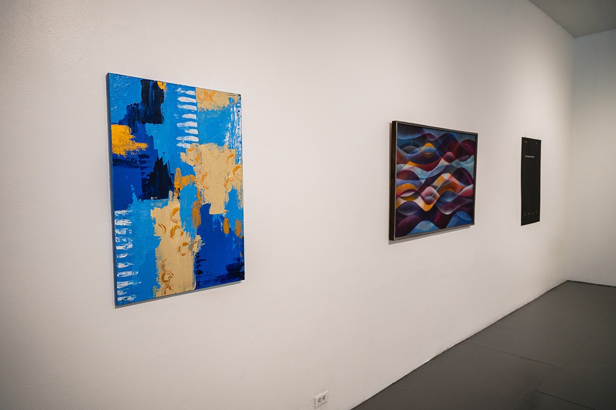 a blue and yellow abstract painting hangs on a white wall other works hang on the same wall in the background.