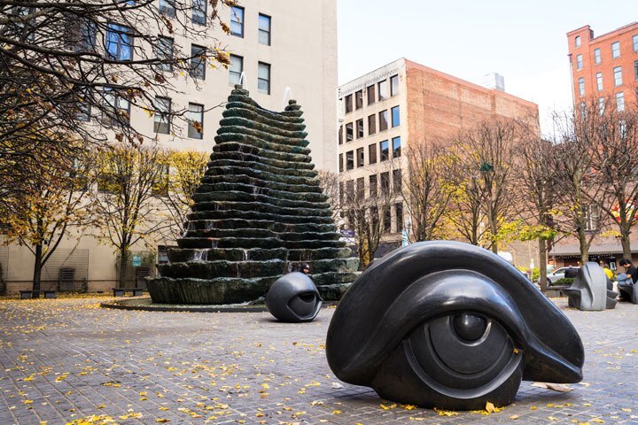 A plaza with leaves with two dark, eye-ball-shaped sculptures and a tiered fountain.