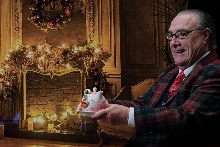 An older man in a red and green plaid jacket holds out a Christmas-themed teapot. In the background, there's a glowing fire and an ornate mantlepiece with golden holiday decorations.