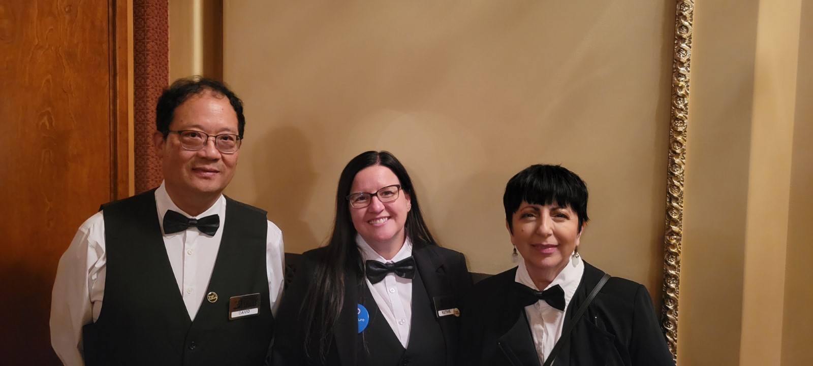 three volunteers in white button downs and black bow ties smile at the camera