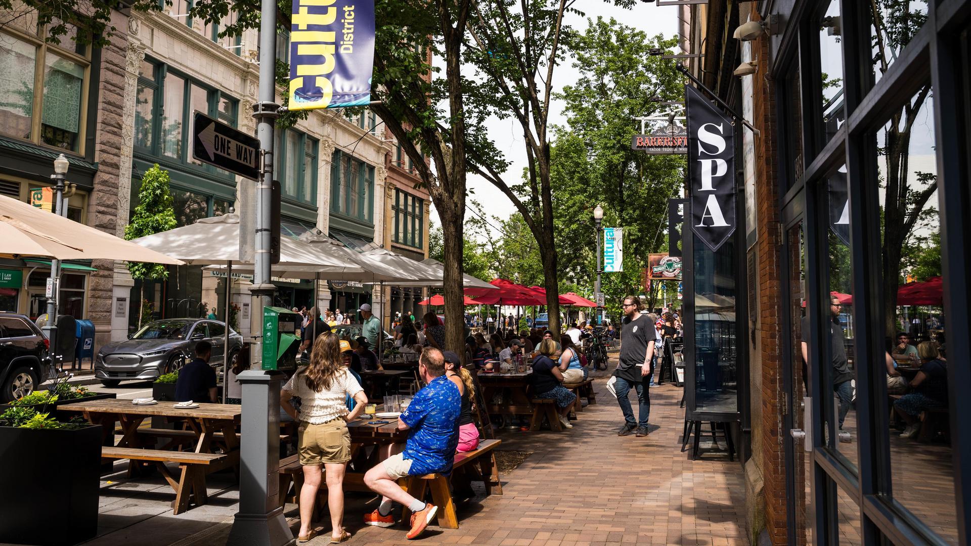 People sit at outdoor tables along a busy city sidewalk lined with trees, restaurants, umbrellas, and storefronts. Some pedestrians walk by, and banners and signs hang from the buildings.