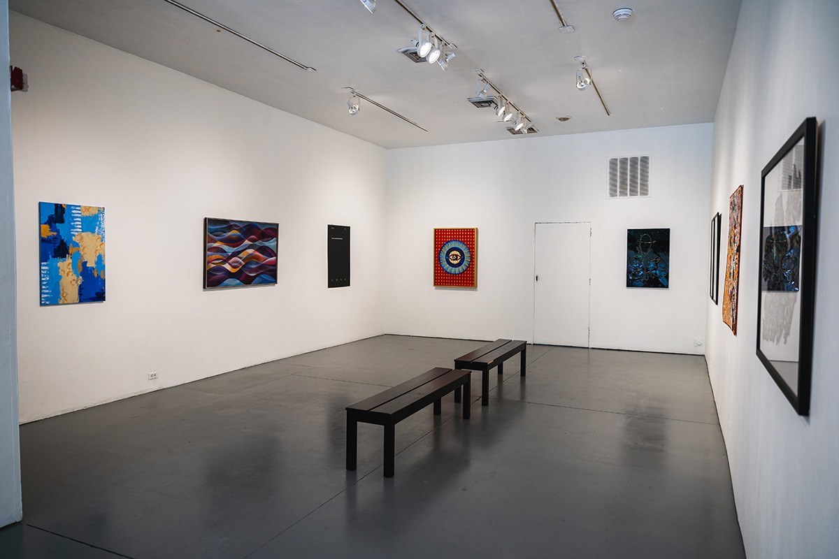 a small gallery space with numerous framed pieces of work hanging on the walls. two black benches sit in the middle of the room