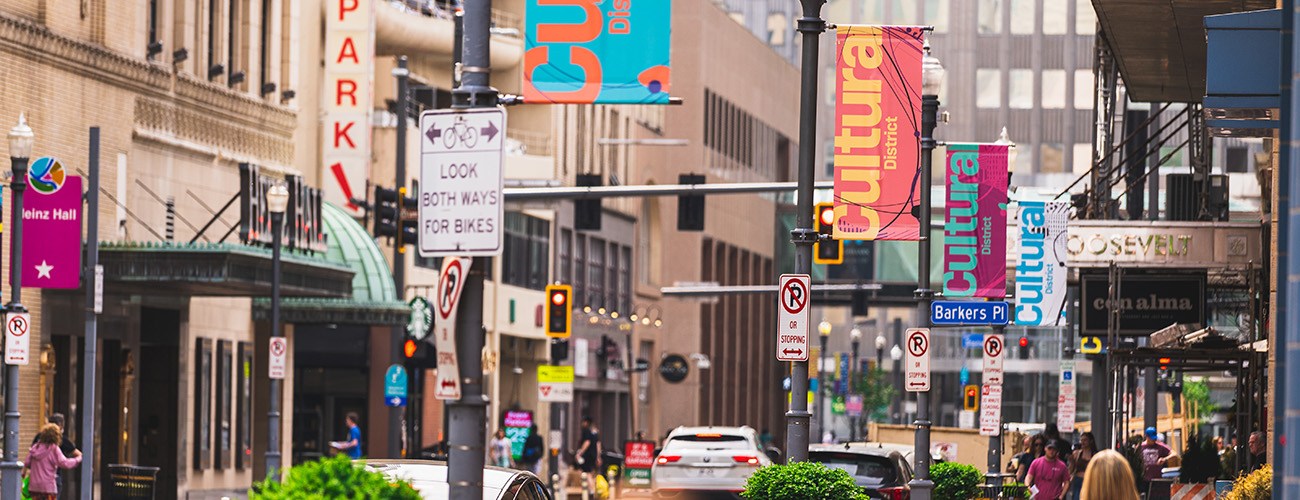 a city block on a sunny day. colorful banners that all say 'cultural district' hang from light poles as people walk past