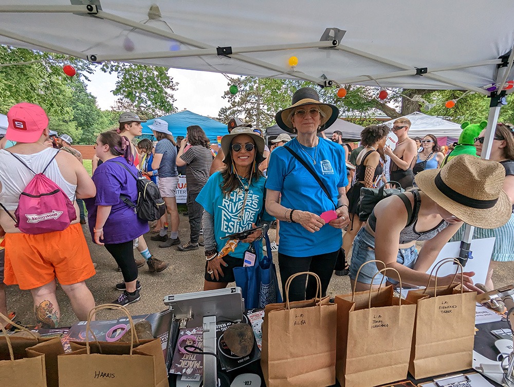 a busy outdoor popup tent. two women in blue t-shirts smile and look at the camera
