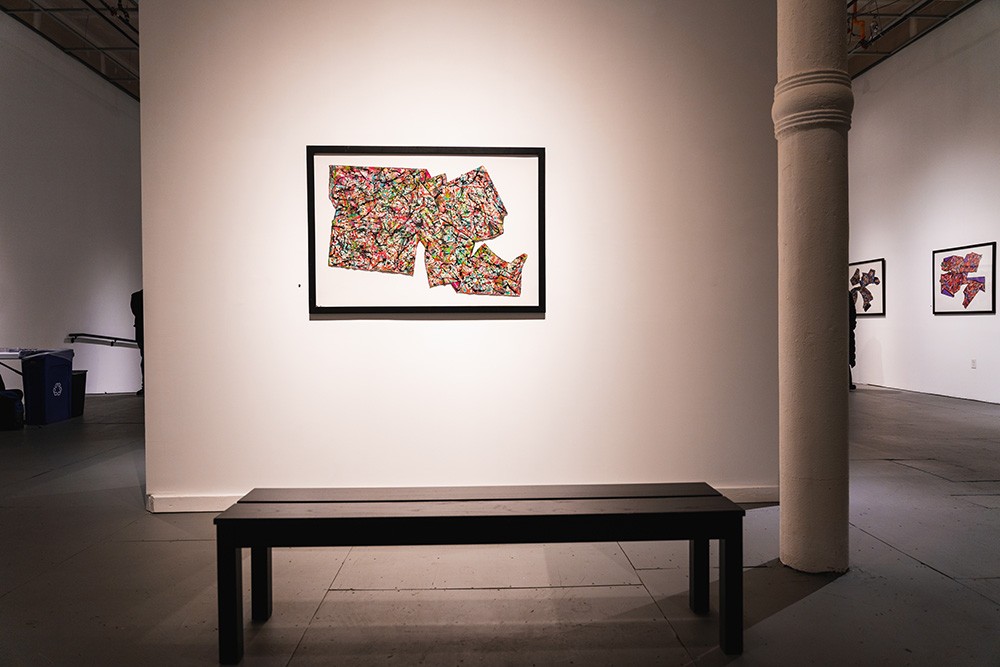 a multicolored mixed media piece hangs on a wall. a black bench sits in front of the piece.