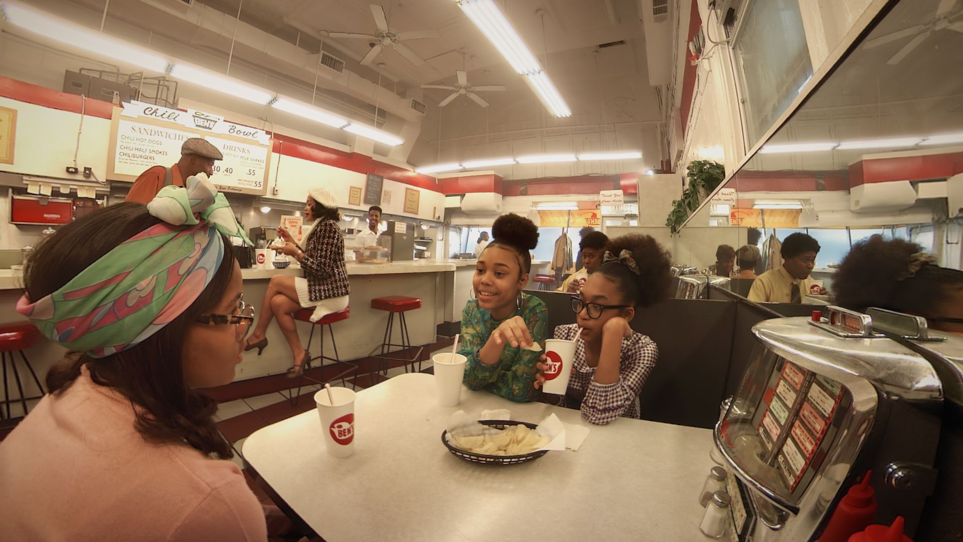 a fisheye point of view image from the perspective of a person sitting in a a restaurant booth, with other people in sixties style clothing milling around the space