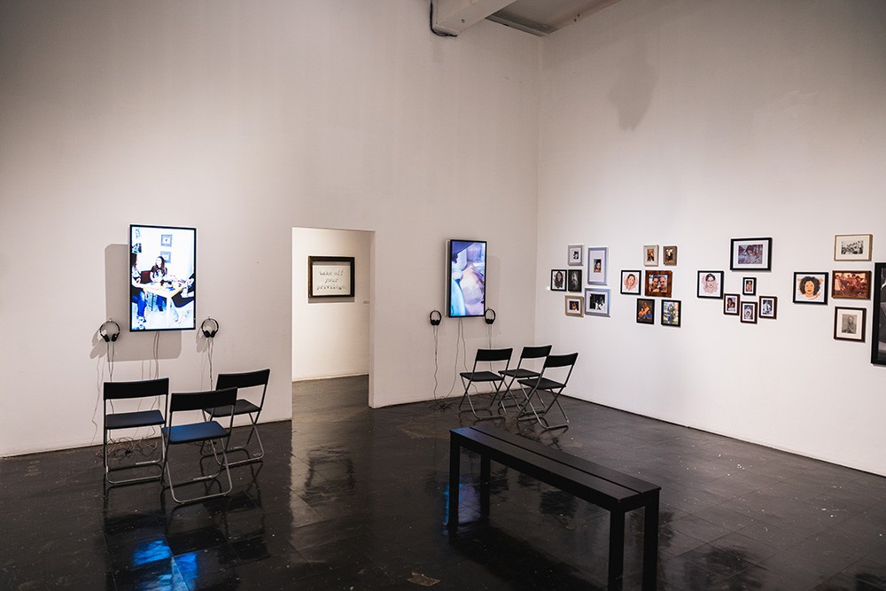 two vertical tv screens hang on a wall with chairs in front of them. smaller photos hang on a perpendicular wall.