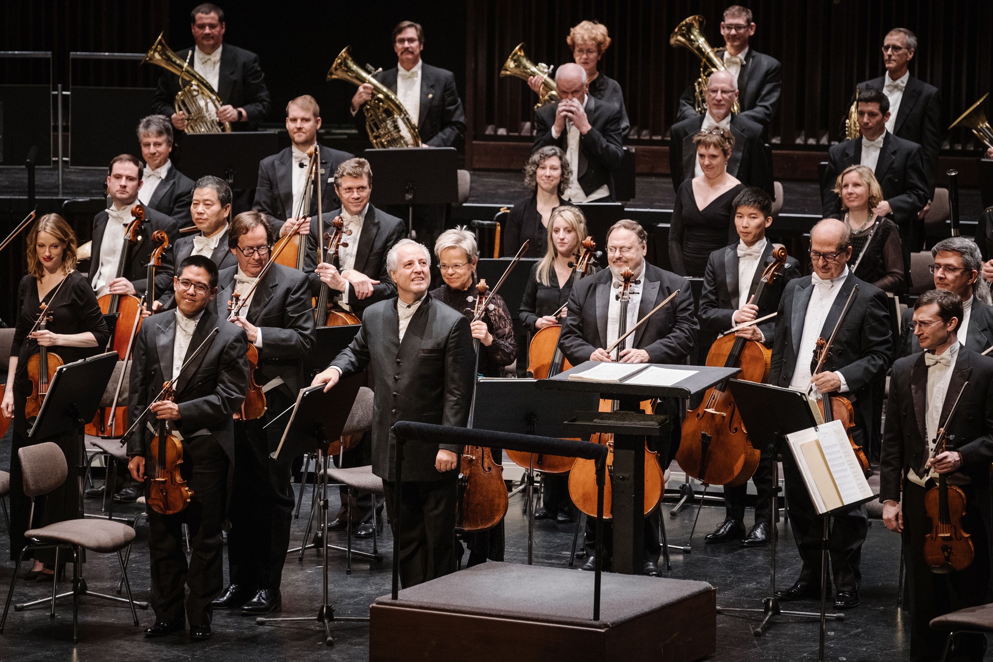 Honeck standing in front of Orchestra holding a baton, all dressed formally