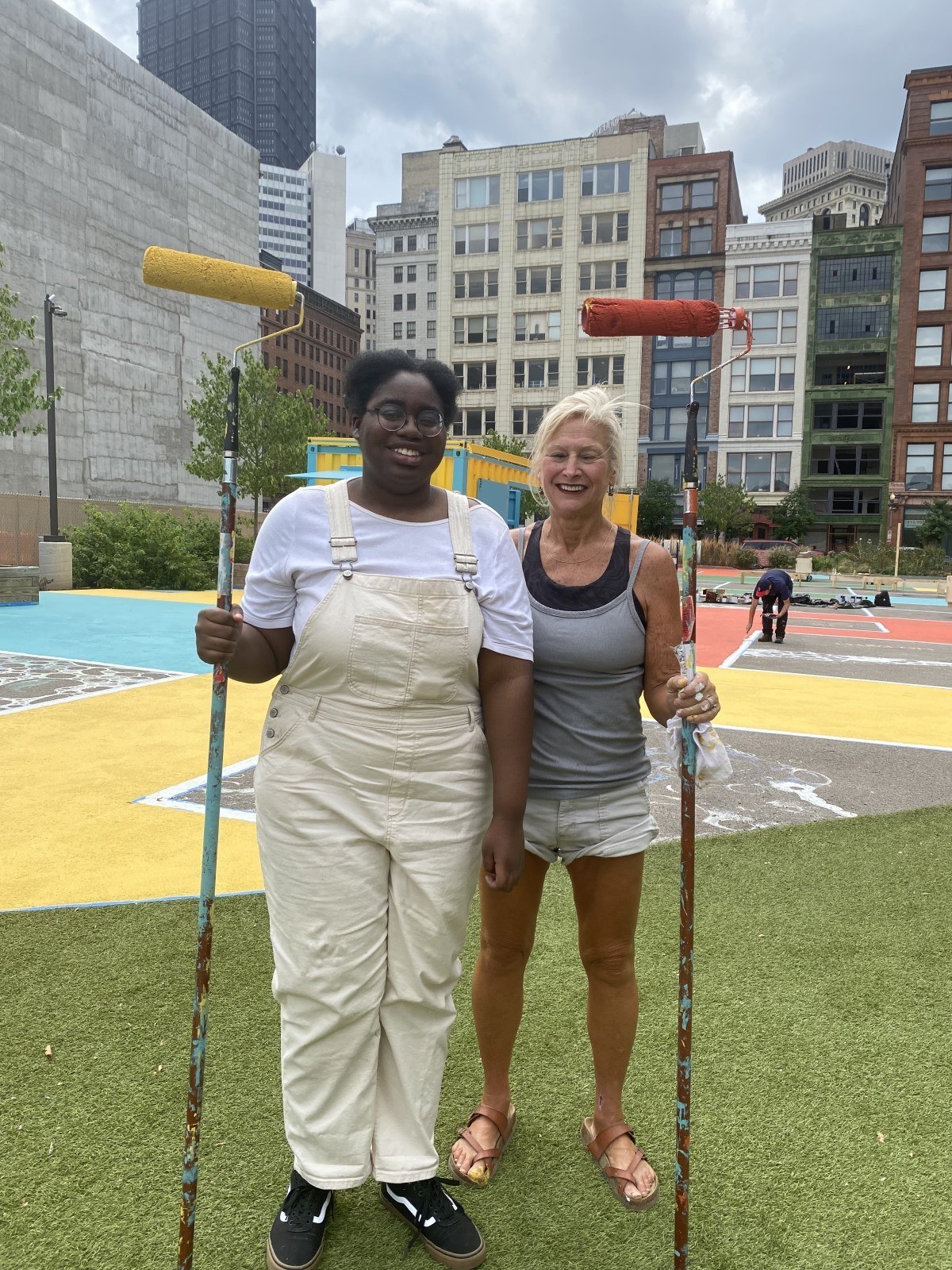 two volunteers hold up paint rollers with a mural on the pavement behind them