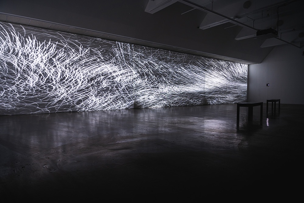 a large, dim gallery space with an abstract pattern of white light projected on a long wall. two benches sit in the middle of the room.
