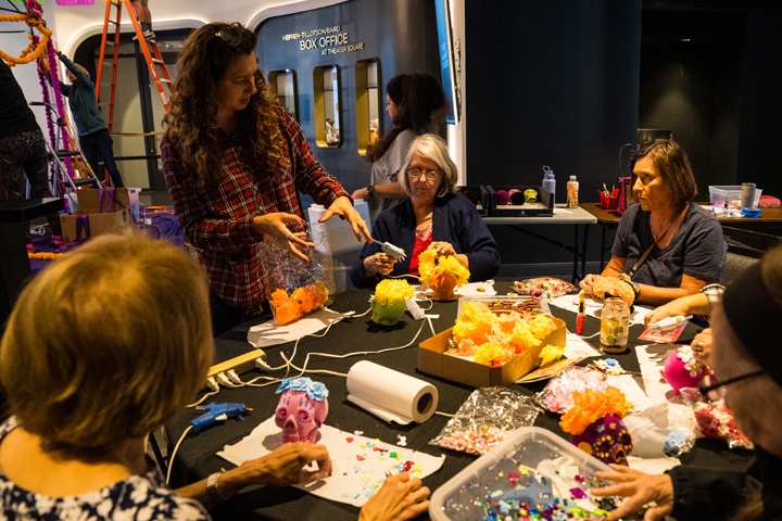 a group sits around a table, decorating colorful skulls with flowers and other jewels.