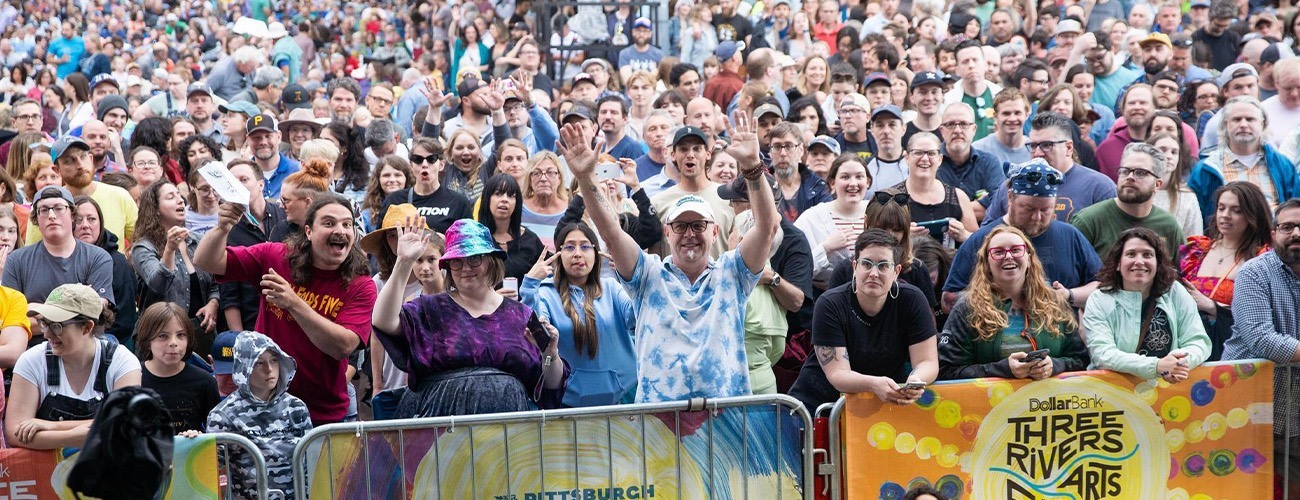 A large, diverse crowd stands outdoors behind barricades at the Three Rivers Arts Festival in Pittsburgh, smiling and waving at the camera on a bright day, with festival banners visible in front.