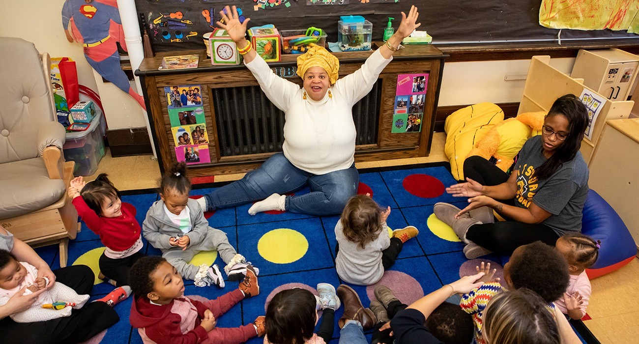 teacher with hands spread out, sitting on ground in front of a group of toddlers