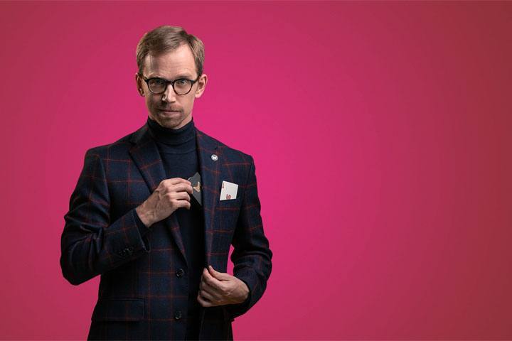 A man stands in front of a hot pink background. He's reaching into his suit jacket for cards.