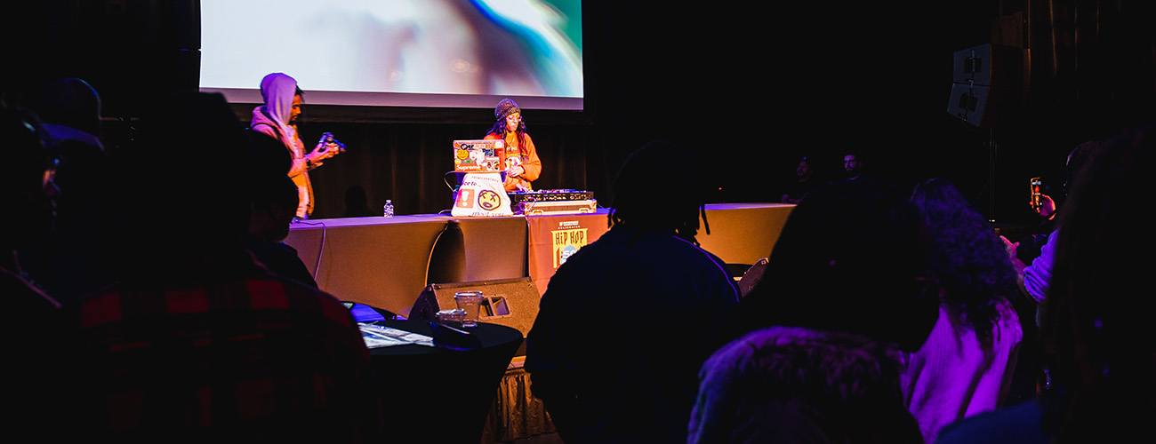 a woman in a red hoodie DJs on stage. silhouettes of people dance in front of the stage.