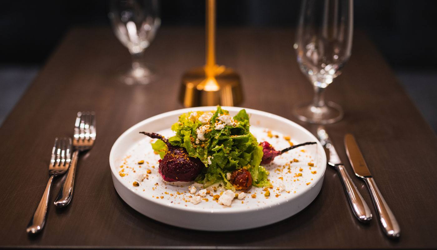 a beet salad on a white plate sits on a dark wood table with silverware