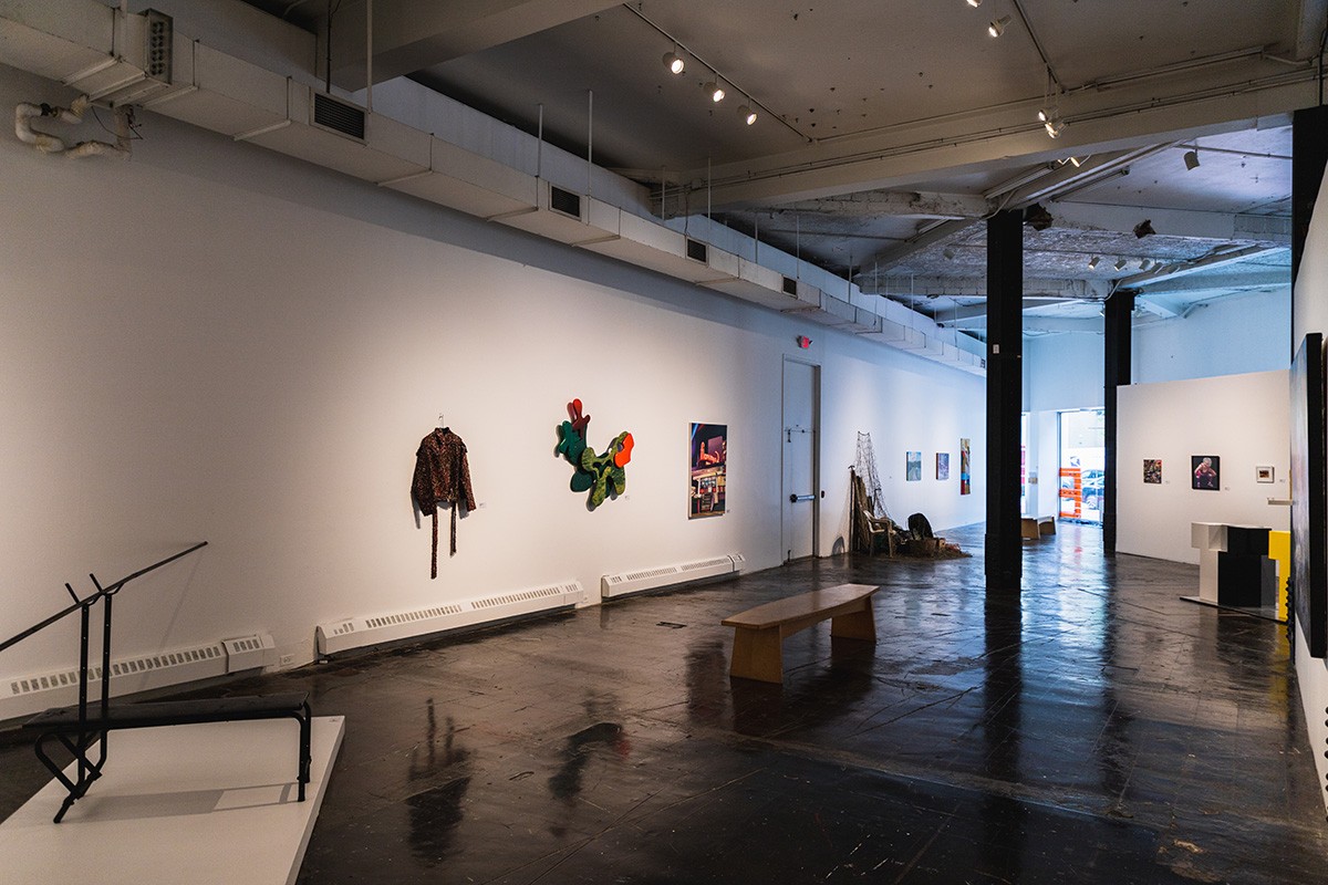 a large industrial gallery space filled with artwork hung on the wall and placed on the floor. a bench sits in the center of the space.