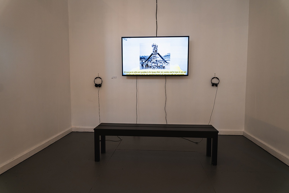 a television screen hanging on a white wall. two sets of headphones hang next to them and a bench sits in front of the display.