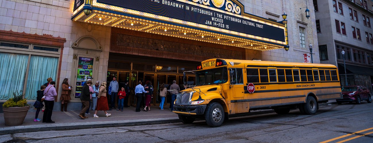 A yellow school bus is parked in front of a theater entrance as a group of people line up outside. The theater marquee above advertises a Broadway event in Pittsburgh.
