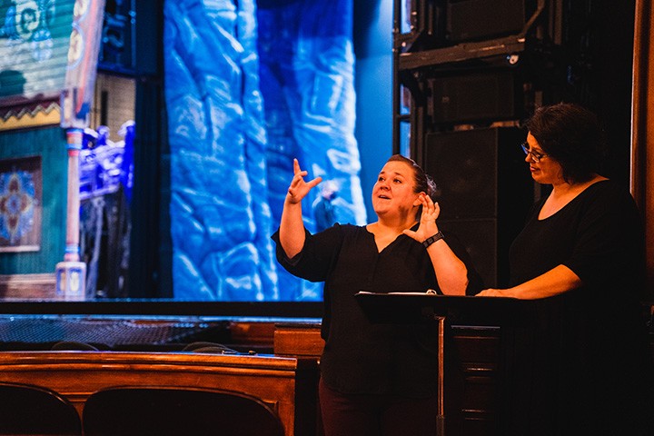 a two white women in black clothing stand in front of a large stage. the woman on the left makes a sign with her hands as the other watches