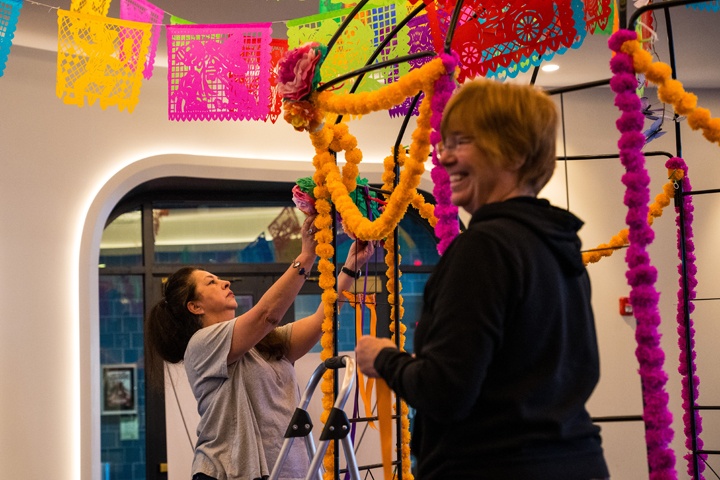 two women stringing golden orange and pink marigolds on the ofrenda.