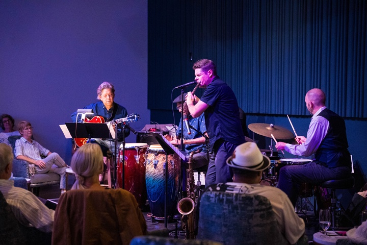 a flute player, two drummers, and a guitarist play in front of a seated crowd.