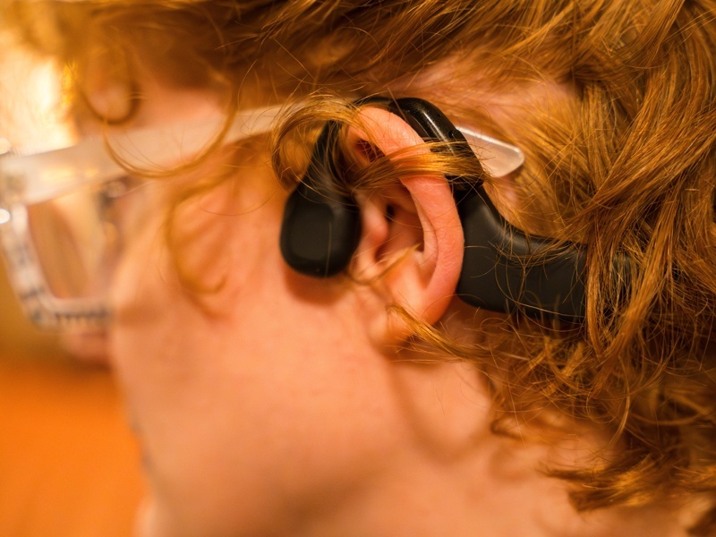 A woman sits in a theater seat wearing glasses, a hearing aid, and bone conduction headphones.