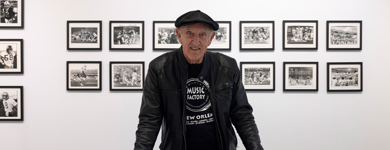 An older man in a black leather jacket and cap stands in front of a wall displaying numerous framed black-and-white photographs. He faces the camera, with his hands resting on a surface in front of him.