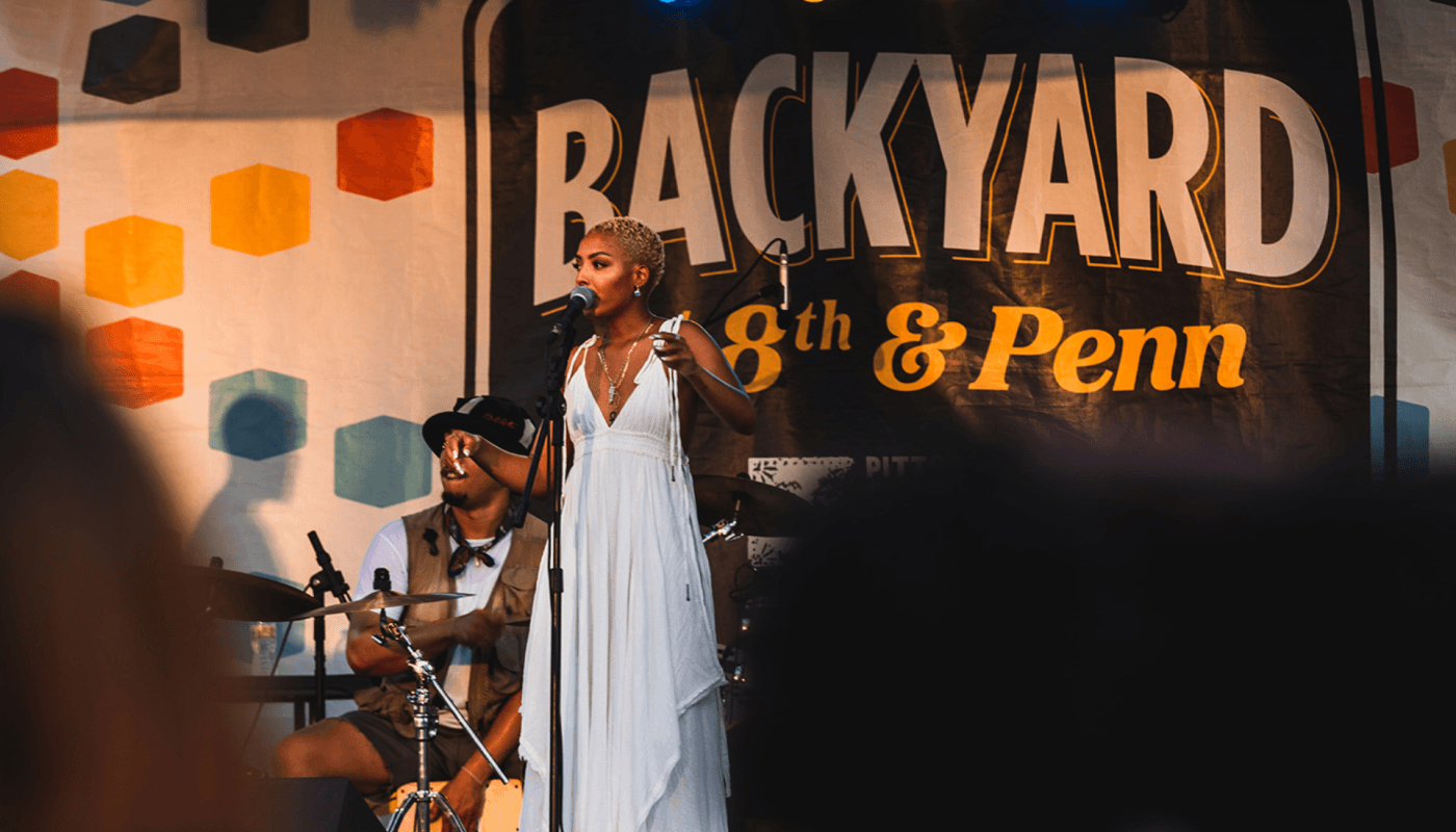 A Black singer performs on stage with the Backyard at Eight and Penn backdrop behind her.